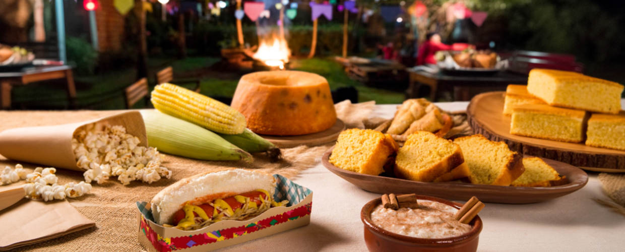 Mesa com bolo de fubá, espiga de milho, pipoca, cachorro-quente, e mais itens de festa junina.
